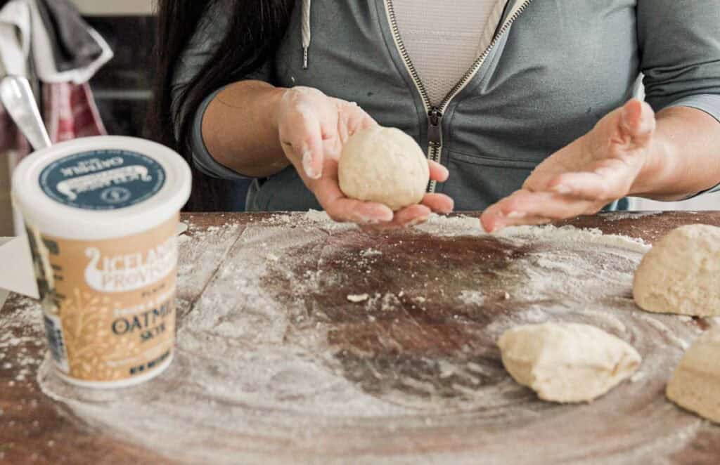 A person shapes dough balls on a floured surface. Nearby, theres a container labeled Icelandic Provisions Oatmilk Skyr. The persons focus is on crafting the dough.